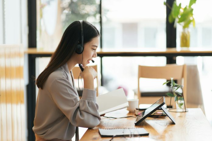 Cheerful beautiful Asian woman wearing headphones and look to digital tablet at video calling meetin