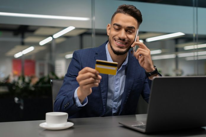 Happy young businessman holding credit card and talking on cellphone