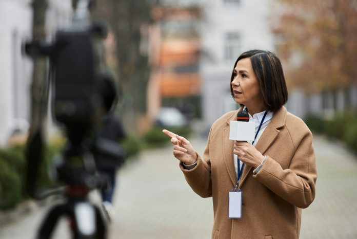 Multiracial female journalist in beige coat reports outdoors during a live news broadcast