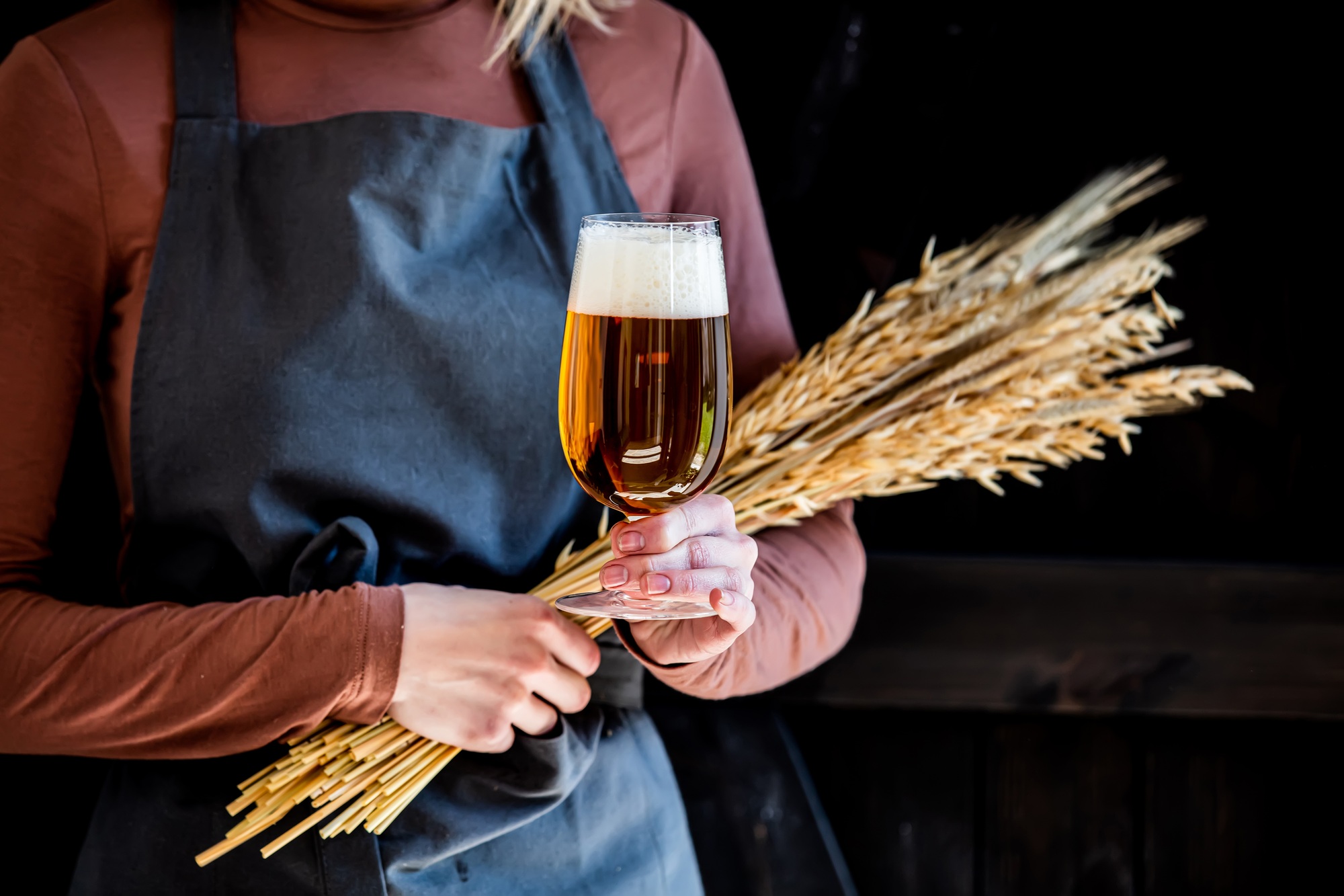 brewer in apron holds glass of beer and wheat spikelets