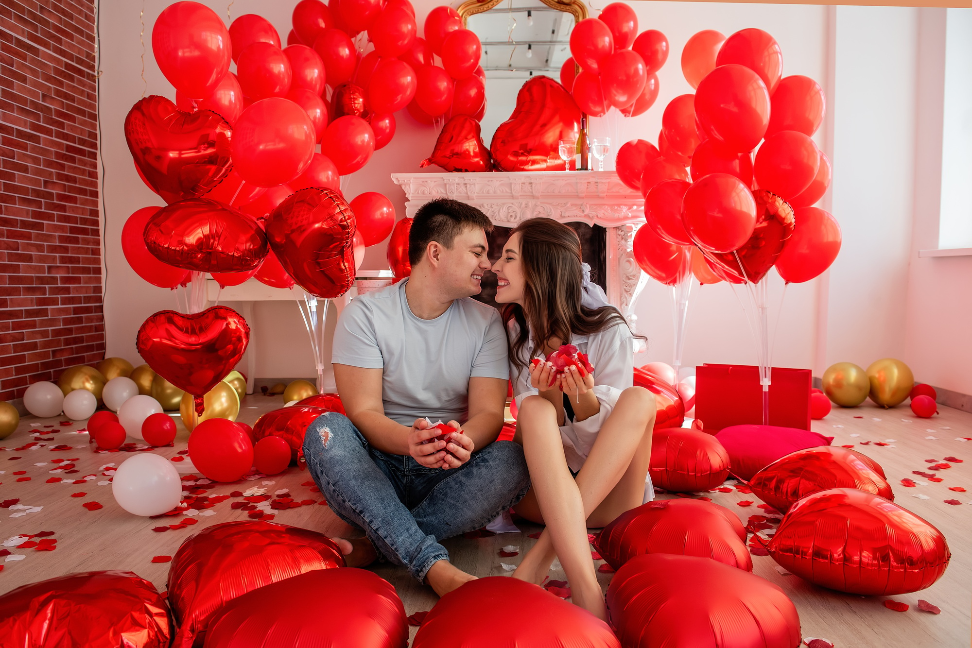 Young man and woman seated close to each other near decorative fireplace adorned with red balloons