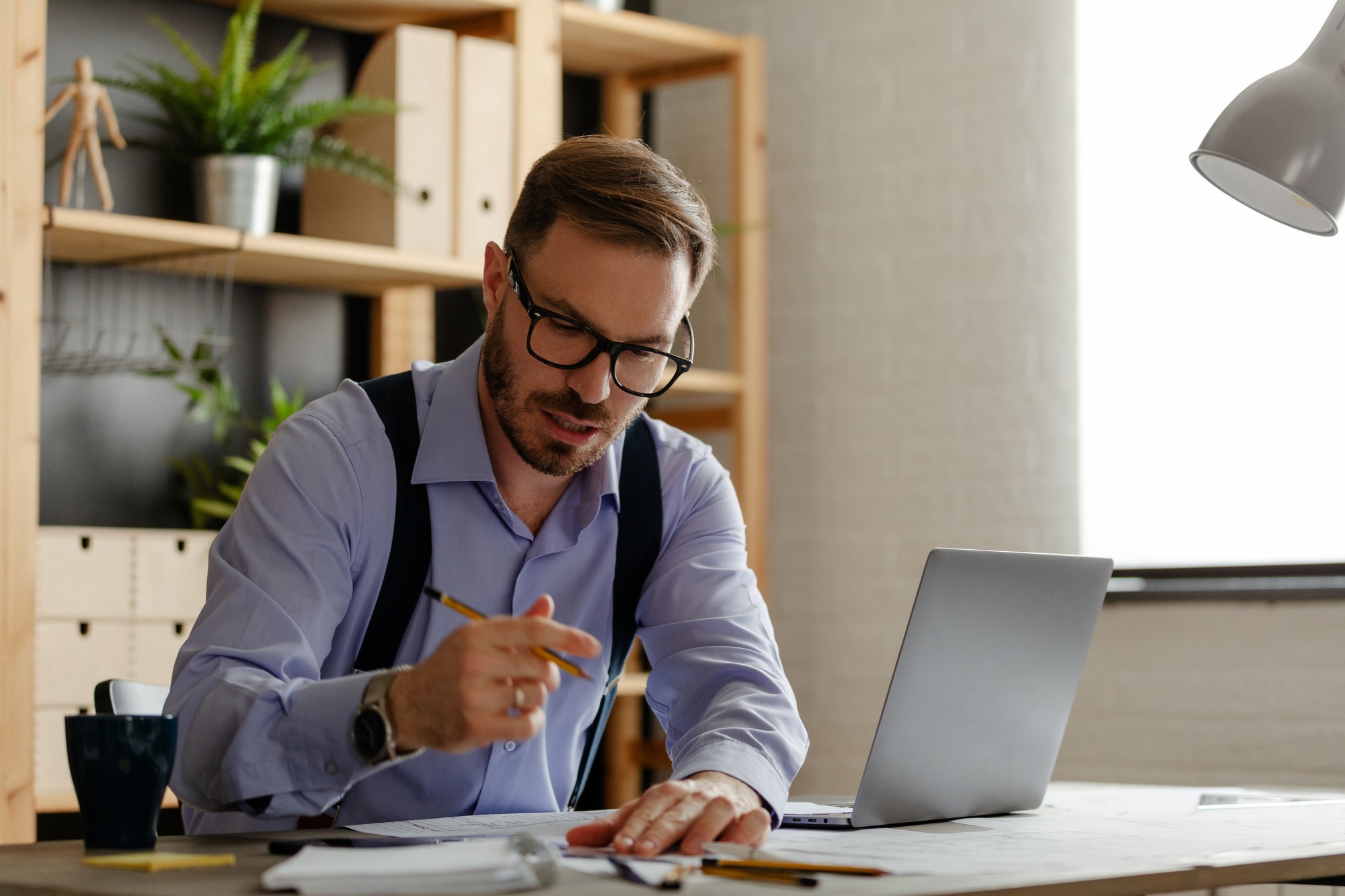 Man with eyeglasses reading information from paper documents and checking in database on laptop