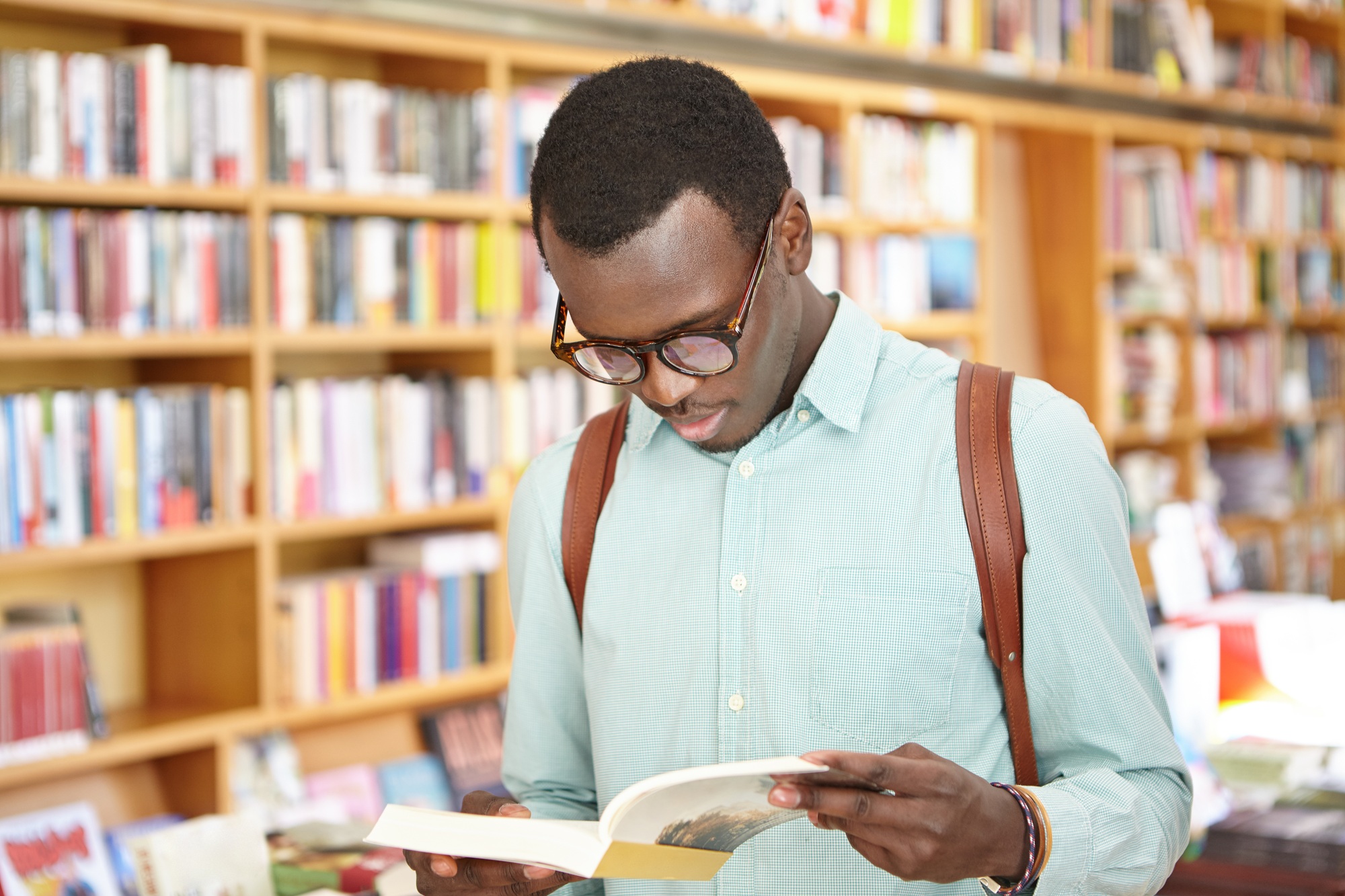 Stylish young African American male in shirt and eyewear looking through book in bookstore standing