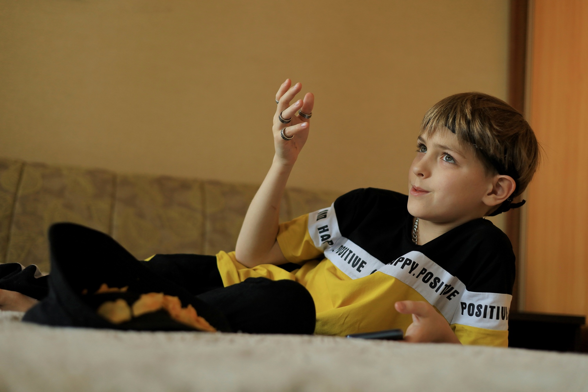 A fashionably dressed boy from generation Z with rings on a hand is lying on the sofa with a phone