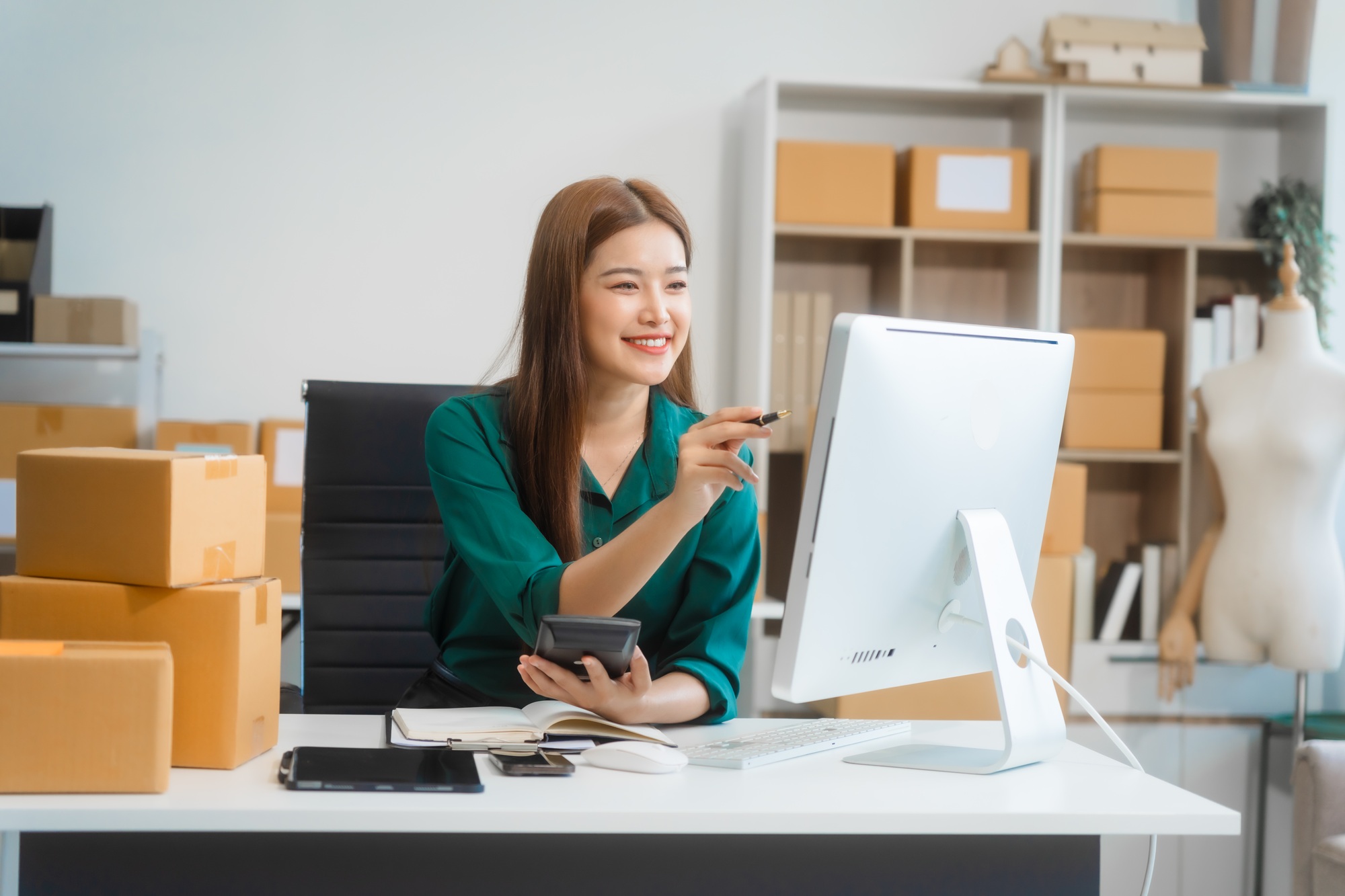 Asian young woman manages her small business, working on a computer amid boxes ready for shipping.