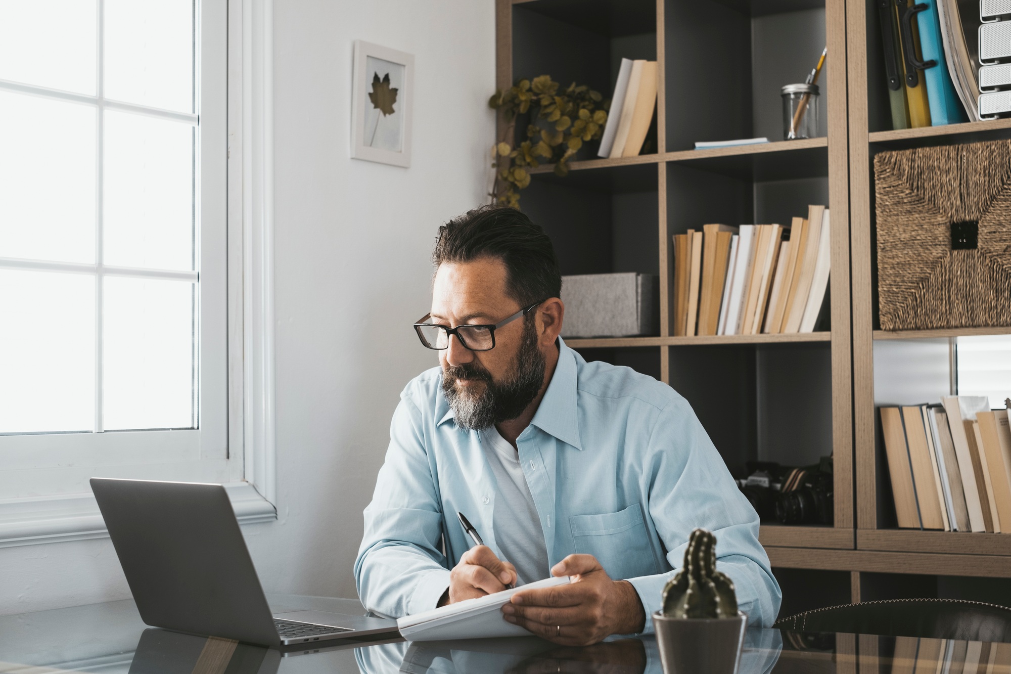 Bearded handsome adult male looking at laptop screen reading email or content and writing notes