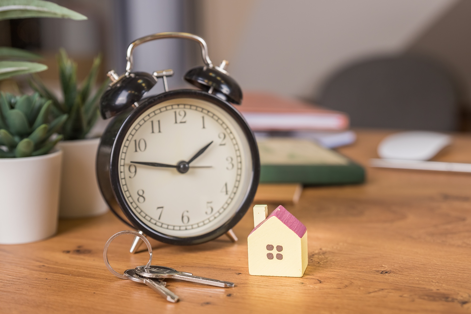 Model house on wooden table, keys, red alarm clock on working place. Real Estate Concept.