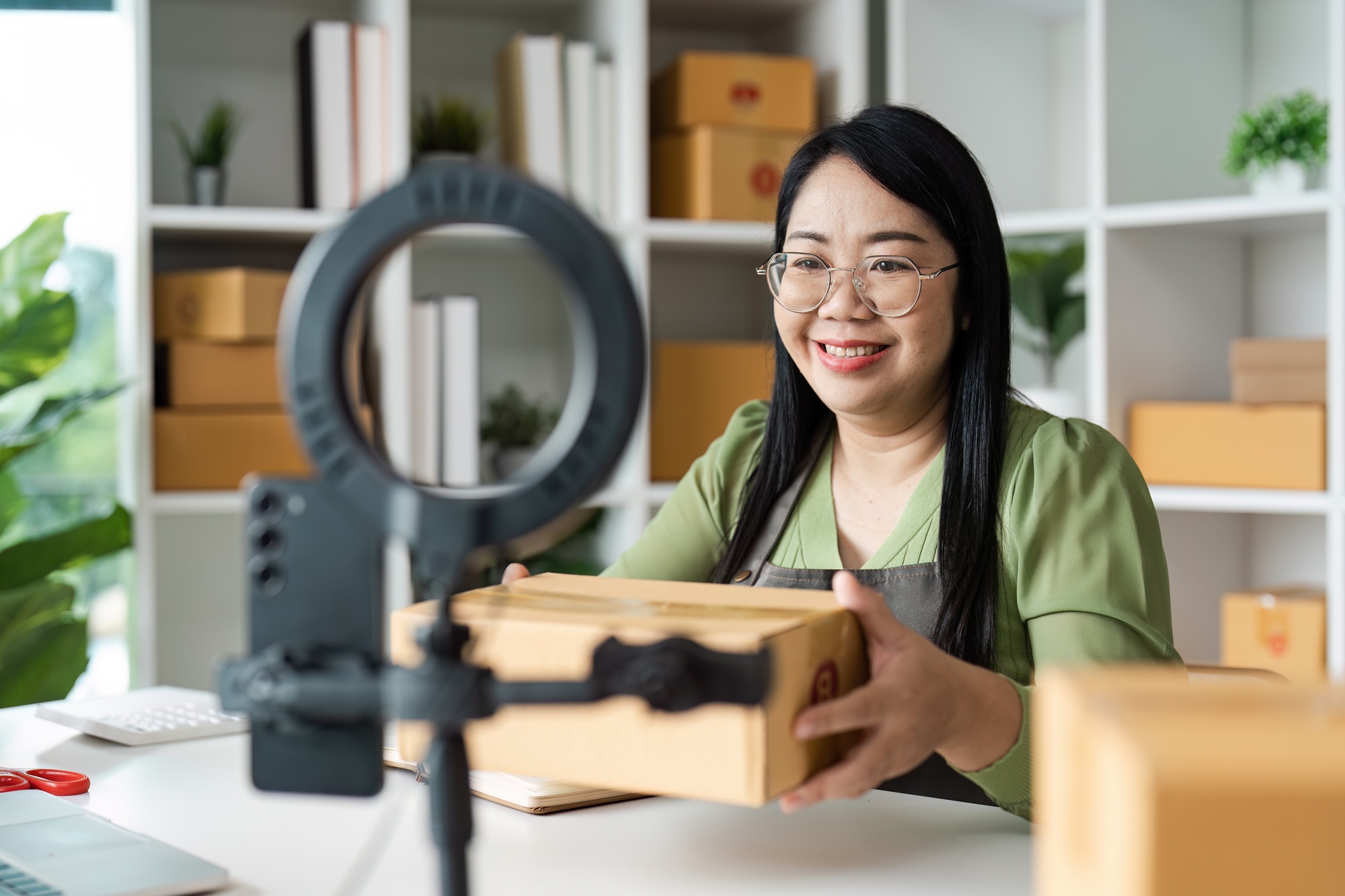 Smiling Woman Conducting Live Selling Online with Packaging Boxes in Modern Home Office Setup