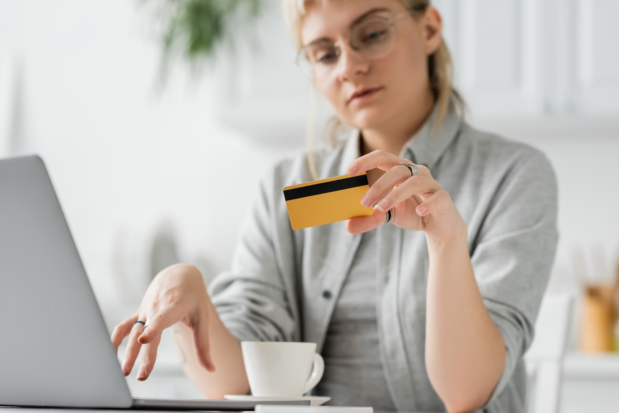 young woman in eyeglasses with tattoo on hand holding credit card, sitting near laptop and cup