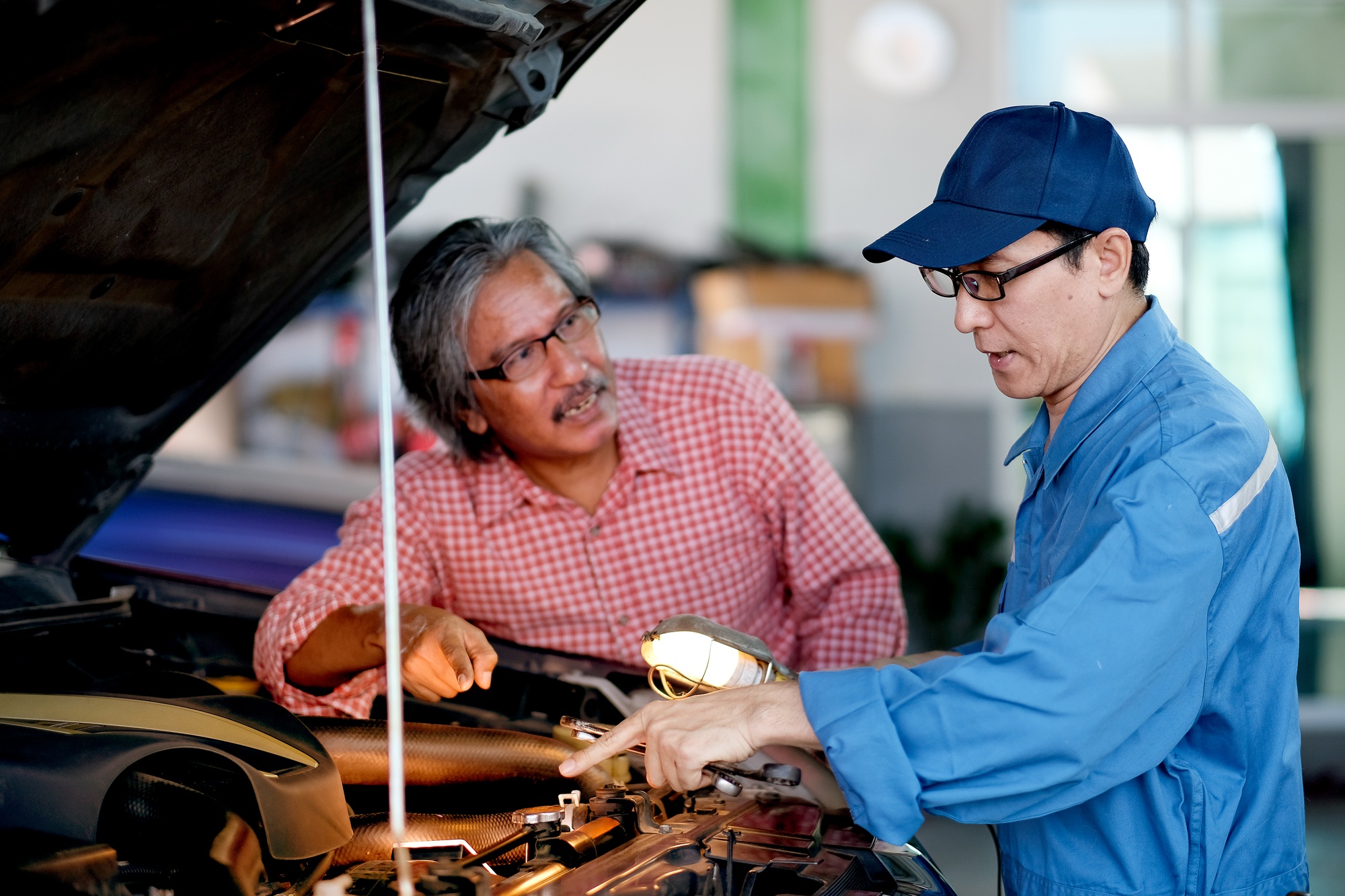 Asian automotive mechanic with blue uniform point to the car engine and explain