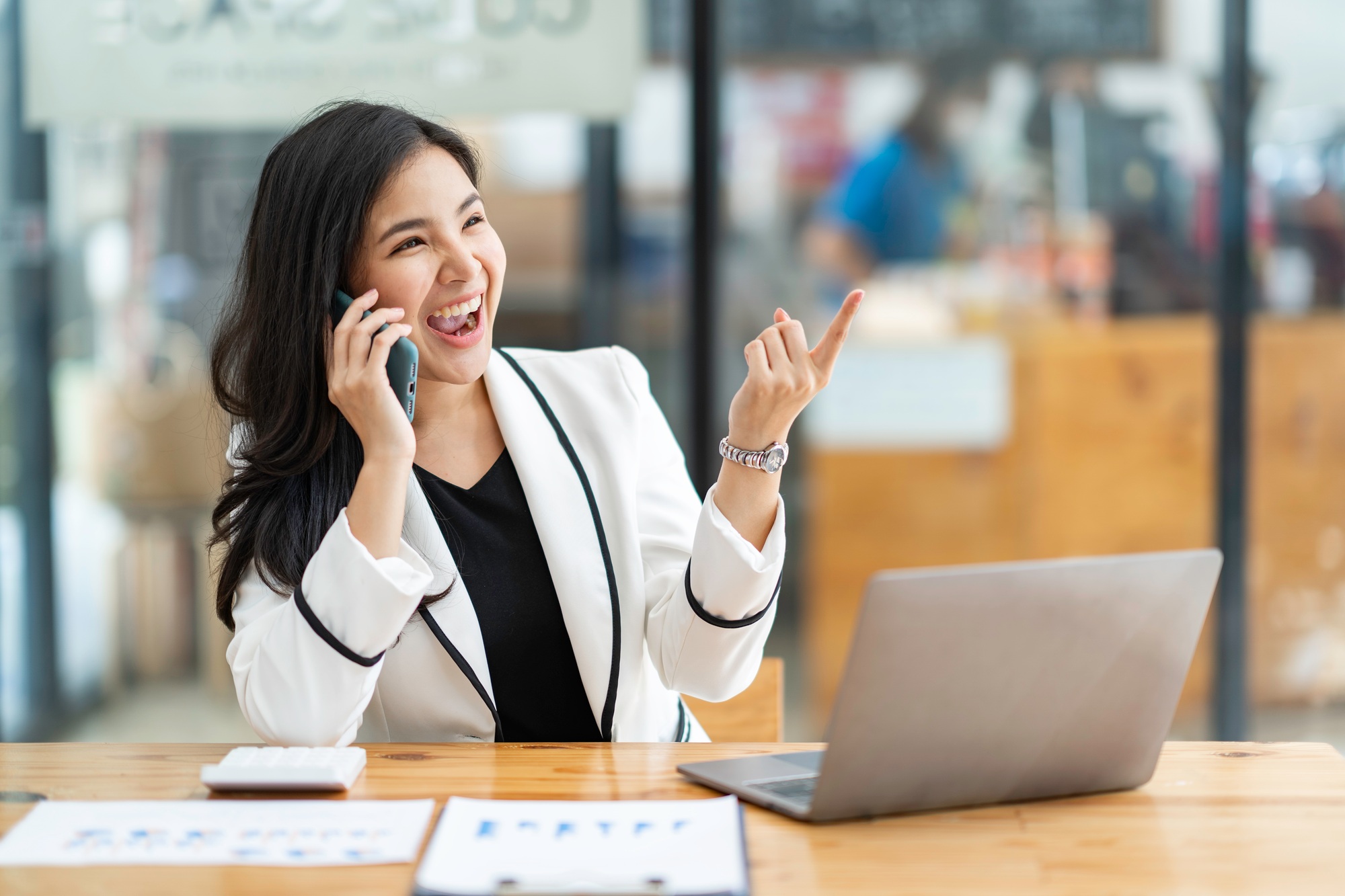 Asian businesswoman happily chatting on the phone with a customer at a table in a modern office.