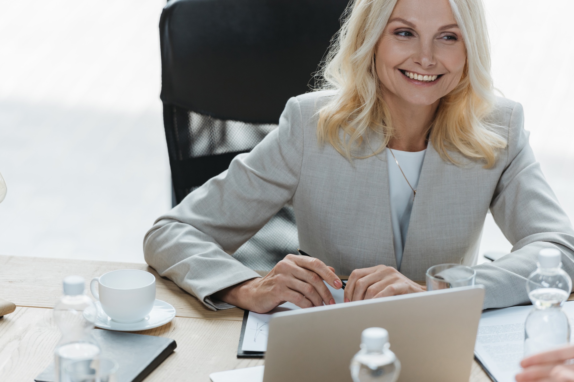 attractive, mature businesswoman smiling while sitting ad desk in meeting room