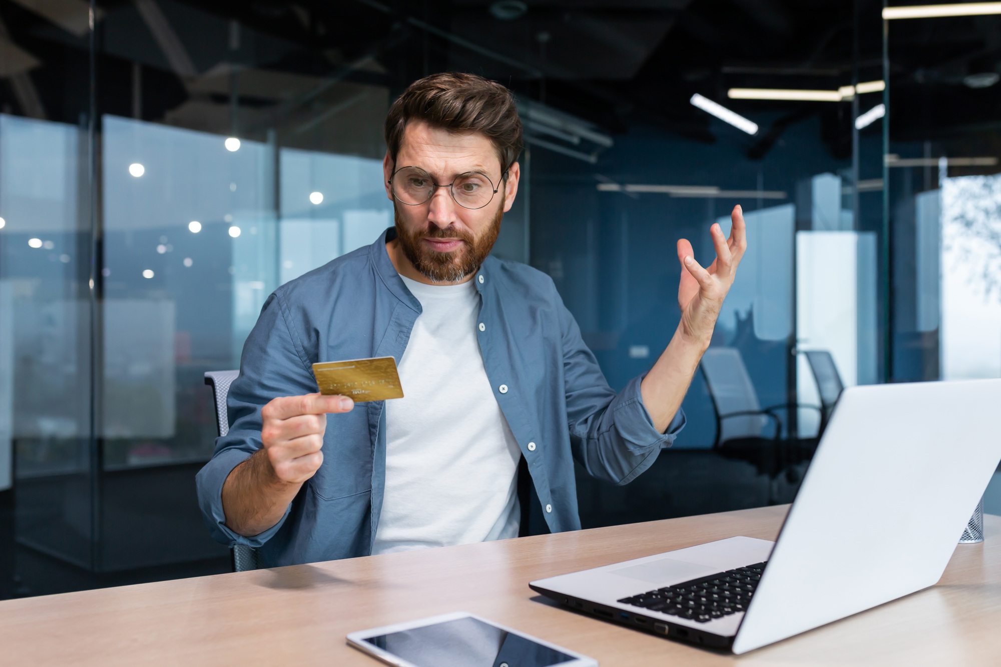 Cheated and disappointed businessman working inside office, man holding bank credit card and trying