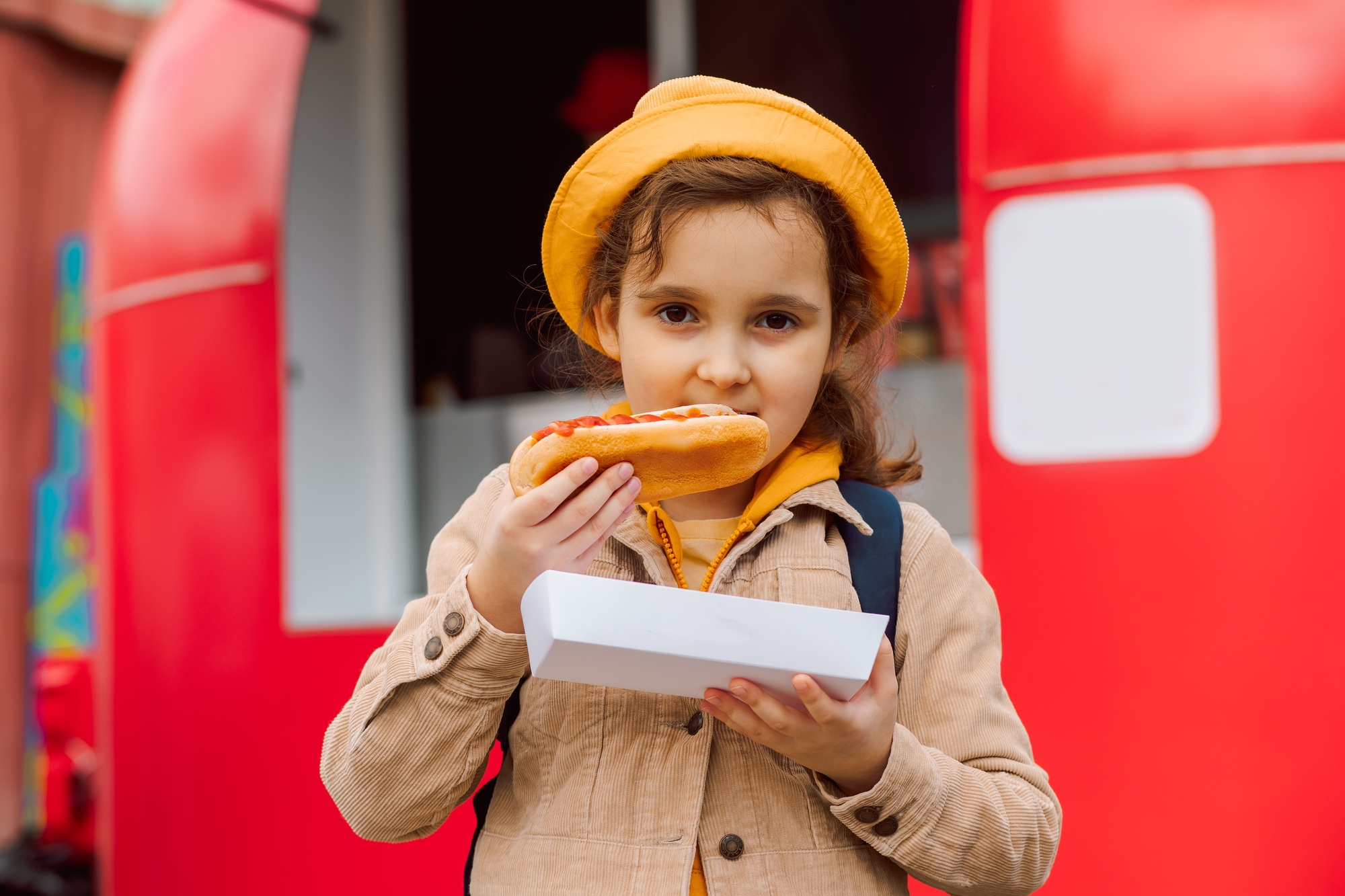 Cute little girl eating hot dog nearby food track on a street. Food delivery or take away food