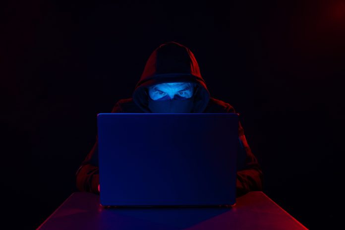 Cyber hacker in hood behind laptop on dark background with red blue light, cyber security concept