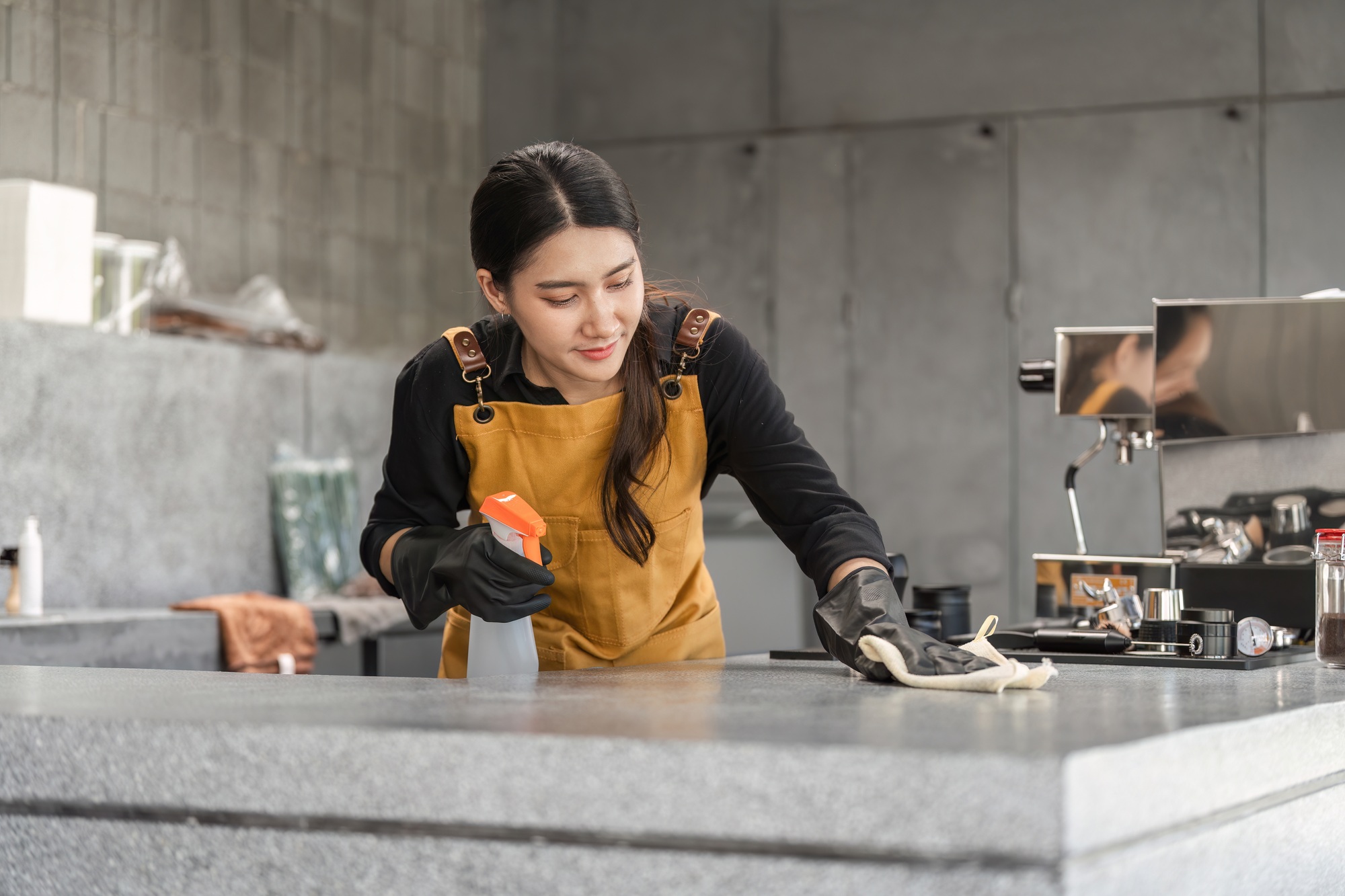 Female Employee Cleaning Modern Coffee Shop Counter with Spray Bottle and Cloth in Industrial Style
