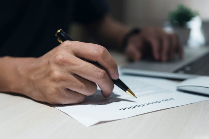 Hand holding pen writing information job description document on desk.