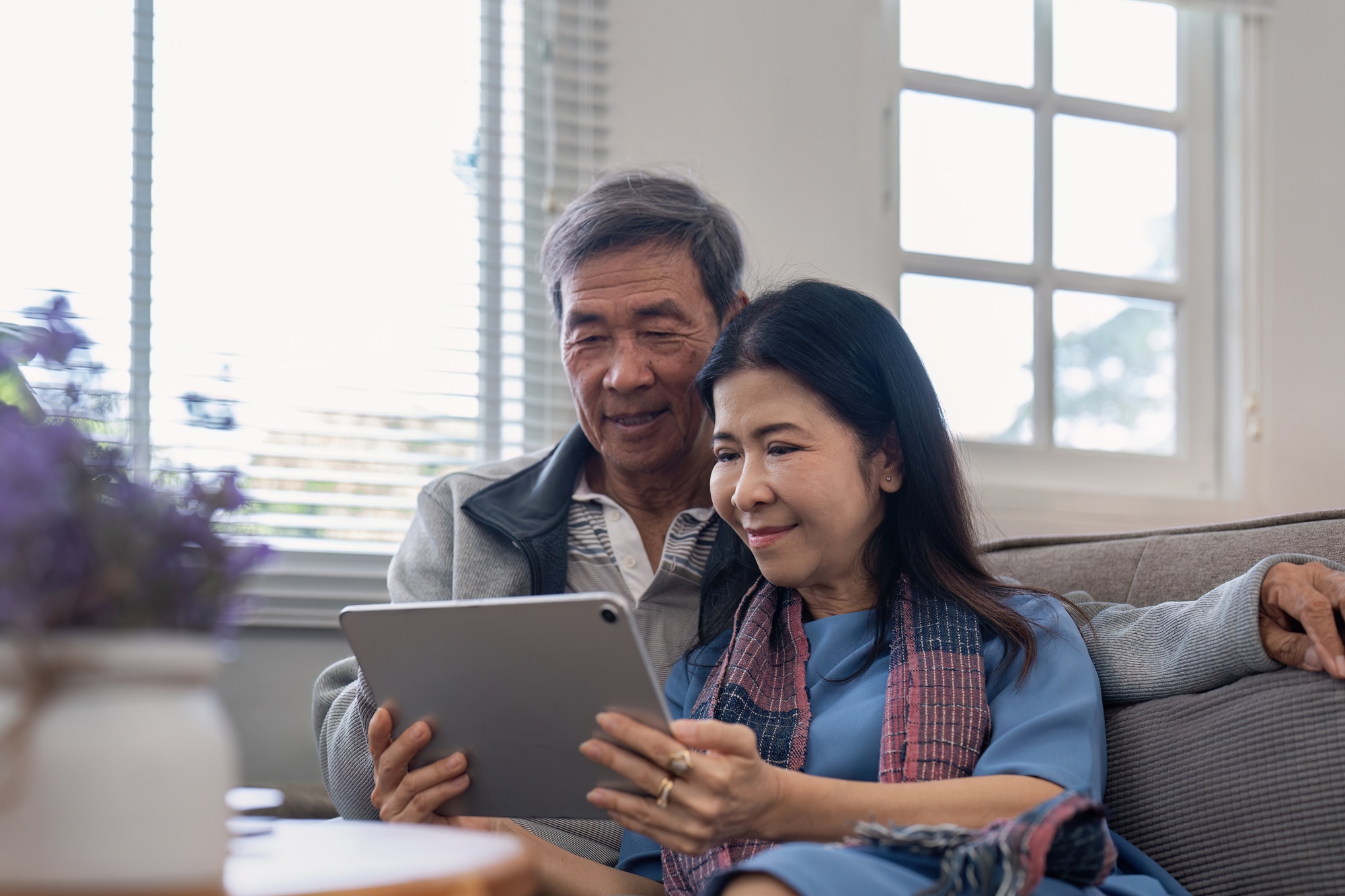 Happy elderly asian couple using tablet sit on sofa doing ecommerce shopping online on website and