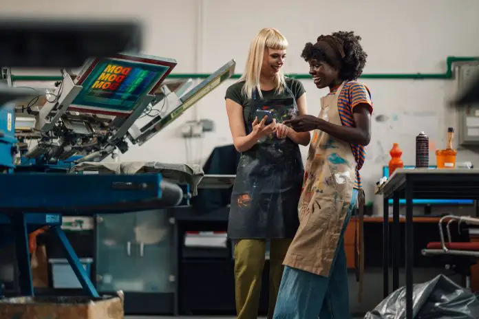 Interracial female graphic technicians smiling at phone at facility.