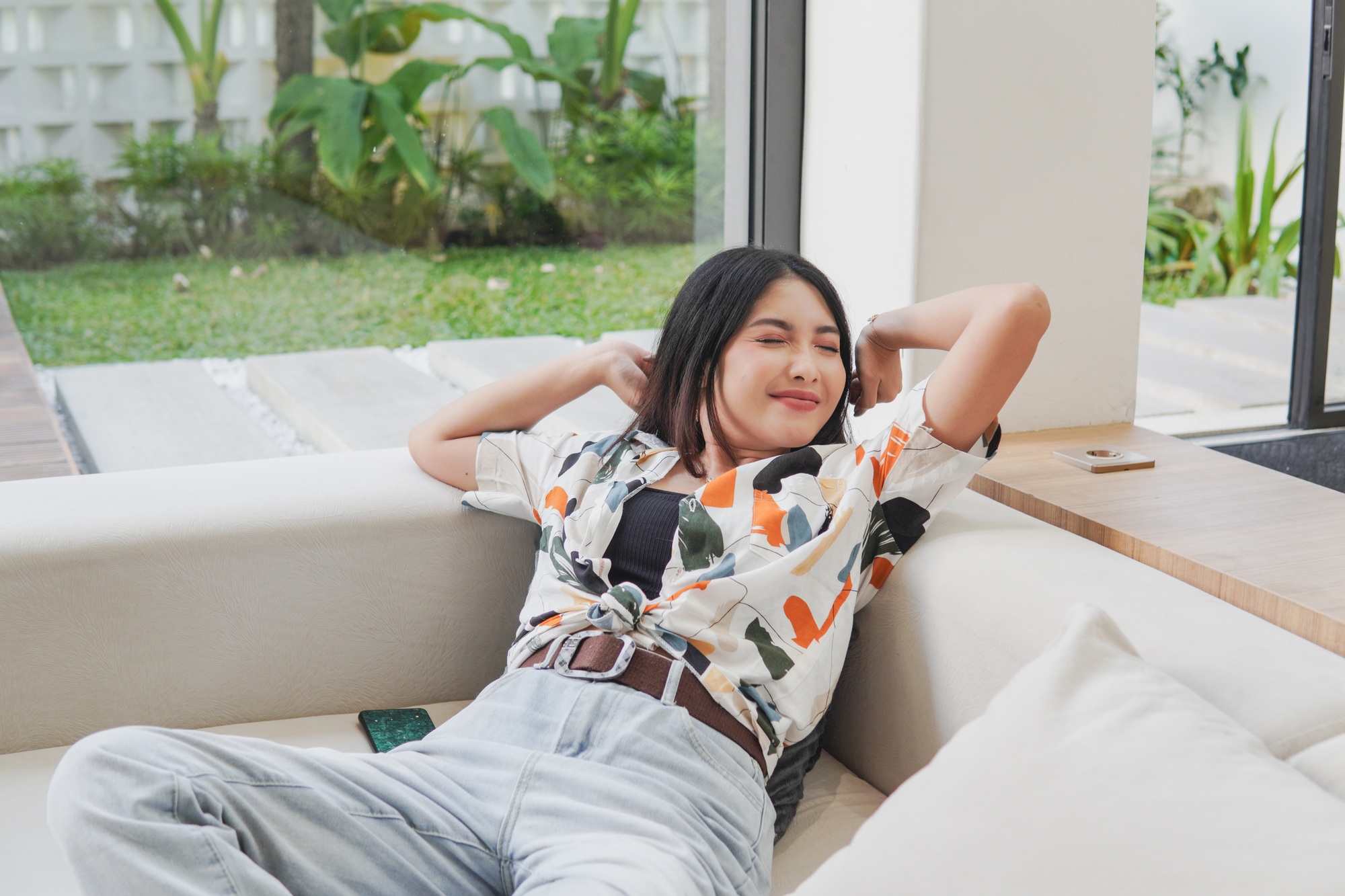 Joyful Asian woman is laying on a couch in a vacation villa with smile and relief expression.