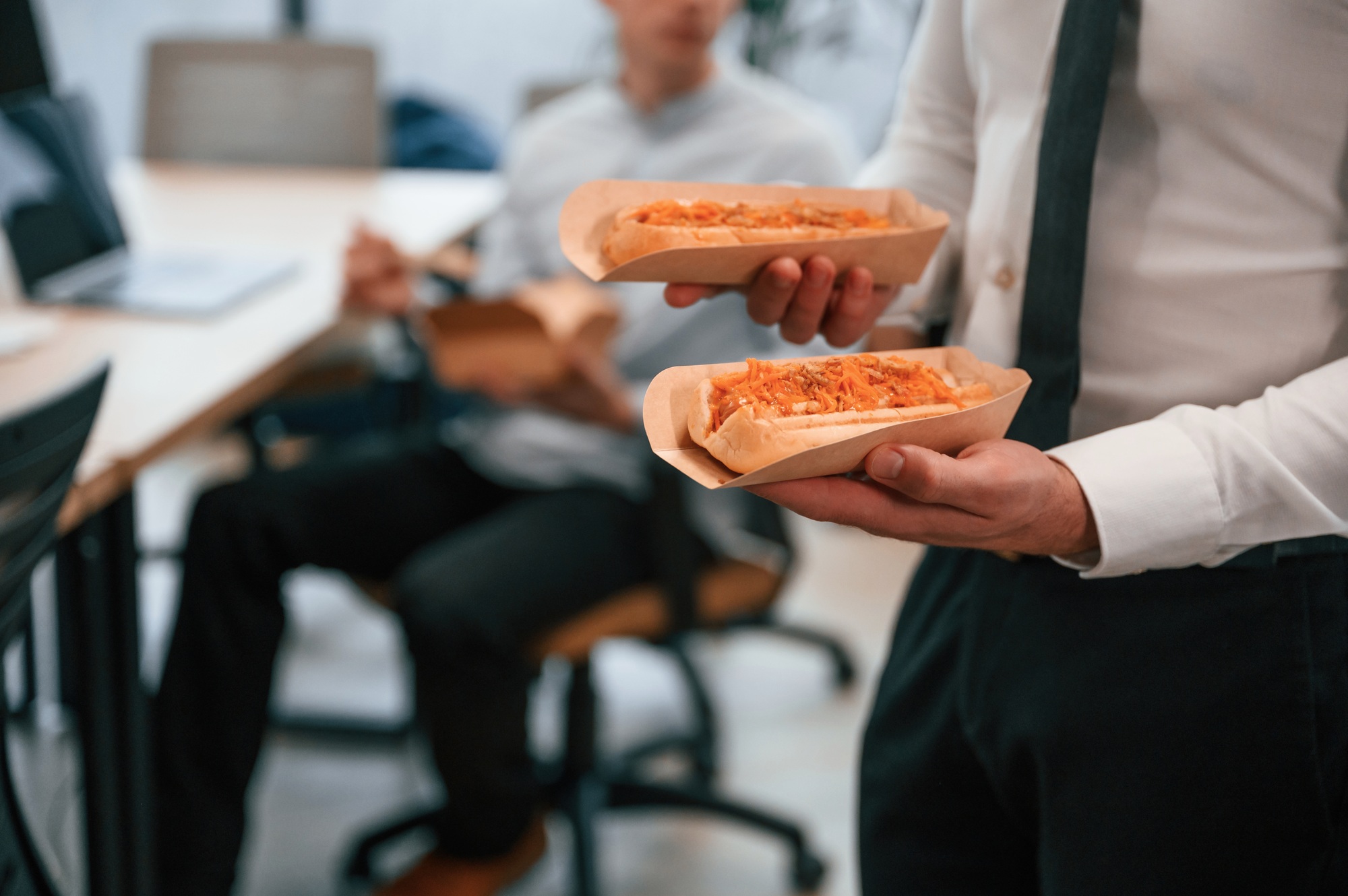 Man in formal wear is in the office, holding hot dogs in hands