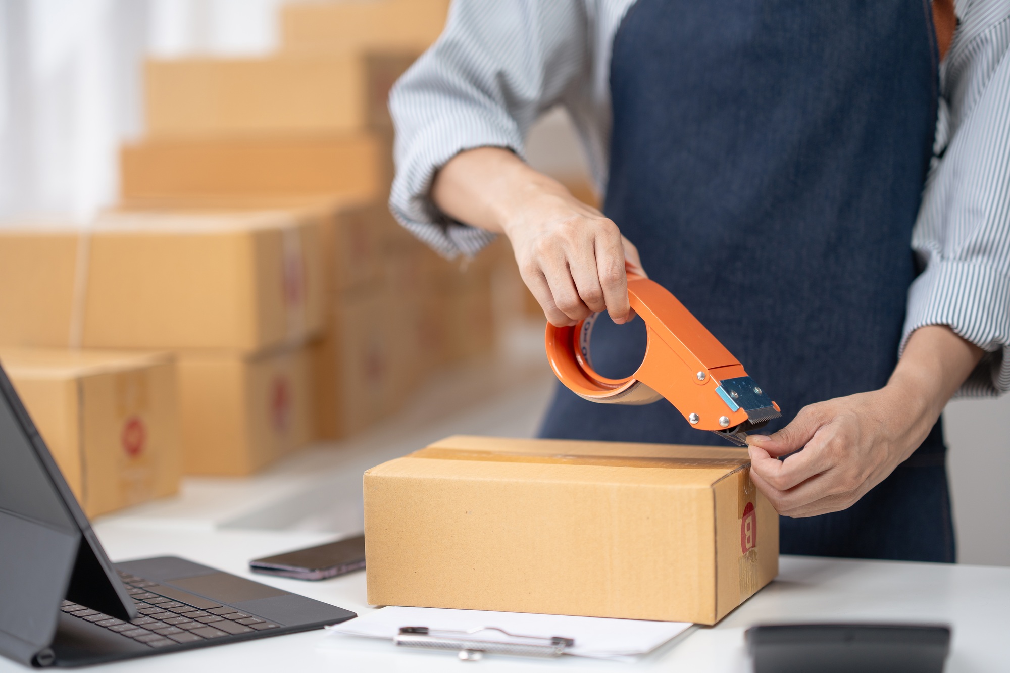 Person sealing a cardboard box with tape in a fulfillment center, surrounded by packages