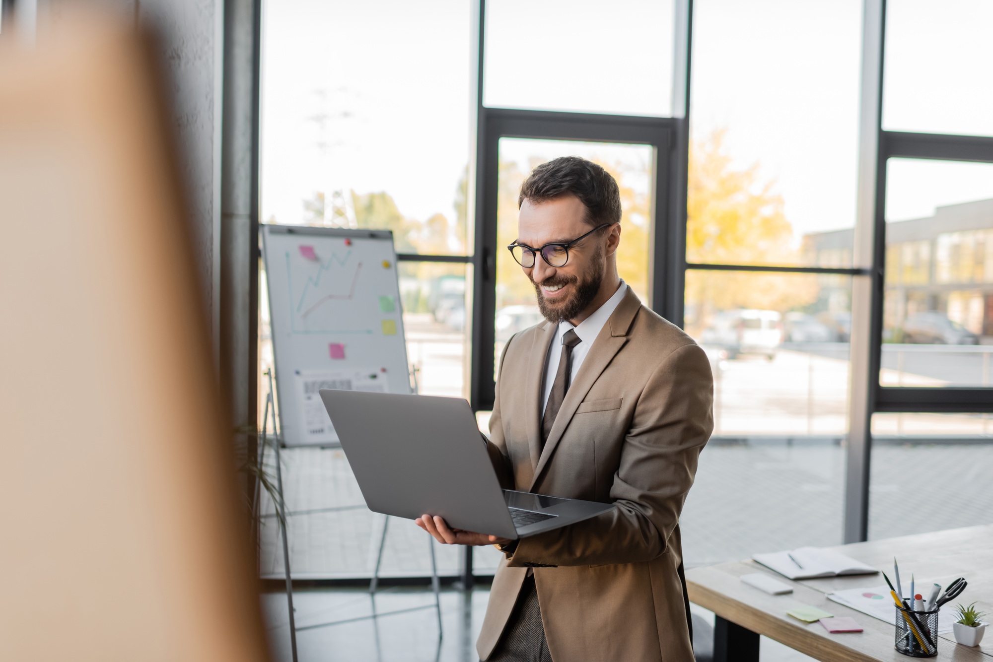 pleased entrepreneur in stylish business attire standing with laptop during video call next to flip