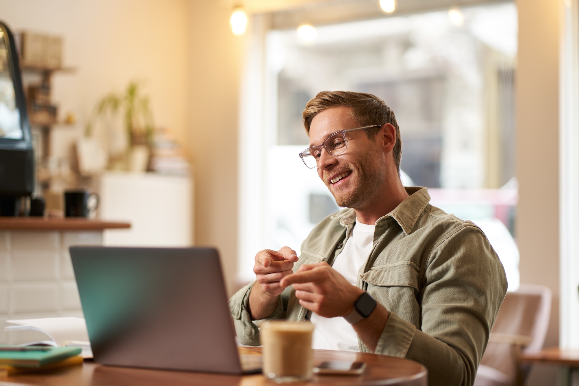 Portrait of handsome, smiling young man, online tutor, businessman working in cafe remotely, waving