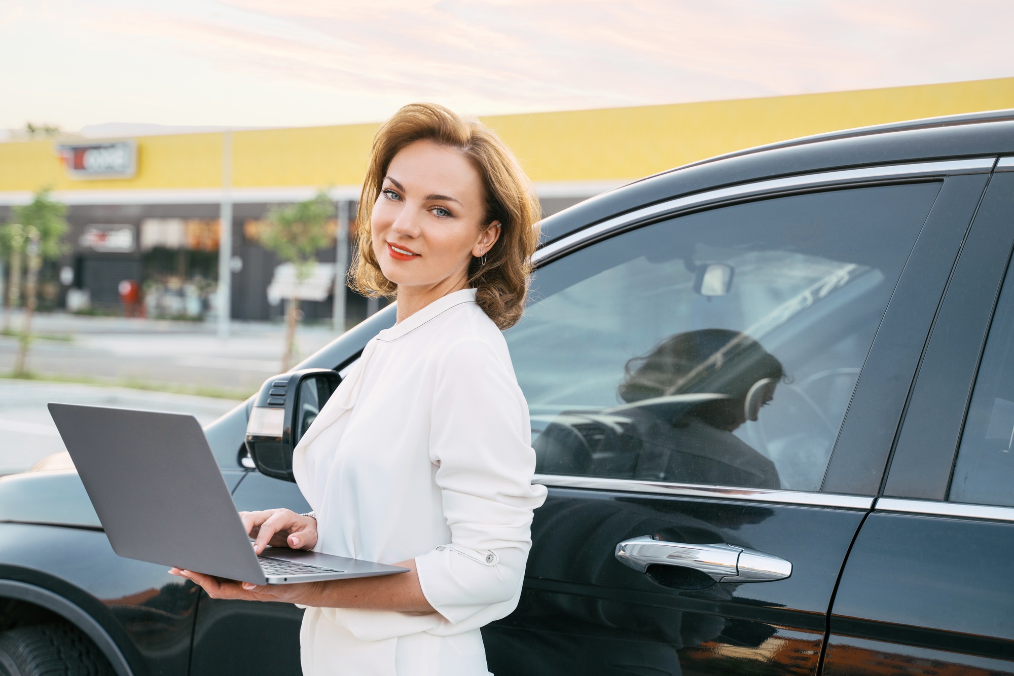 Saleswoman using laptop standing near new car. Dealership, car rental concept