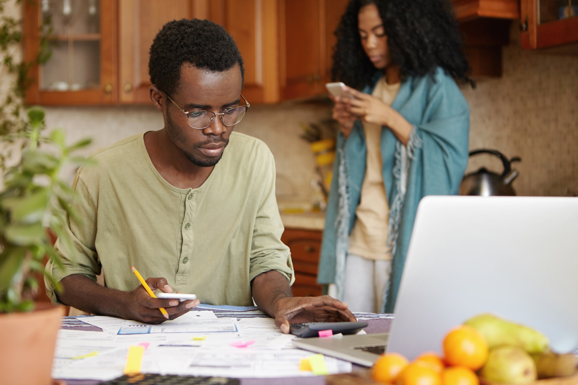 Serious young dark-skinned male in spectacles using cell phone and calculator while calculating fami