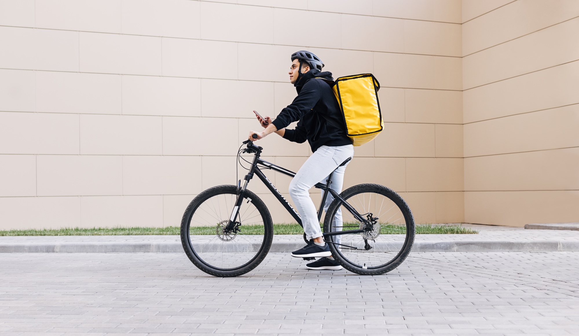 Side view of a young messenger with termal backpack man riding his bike and using mobile phone