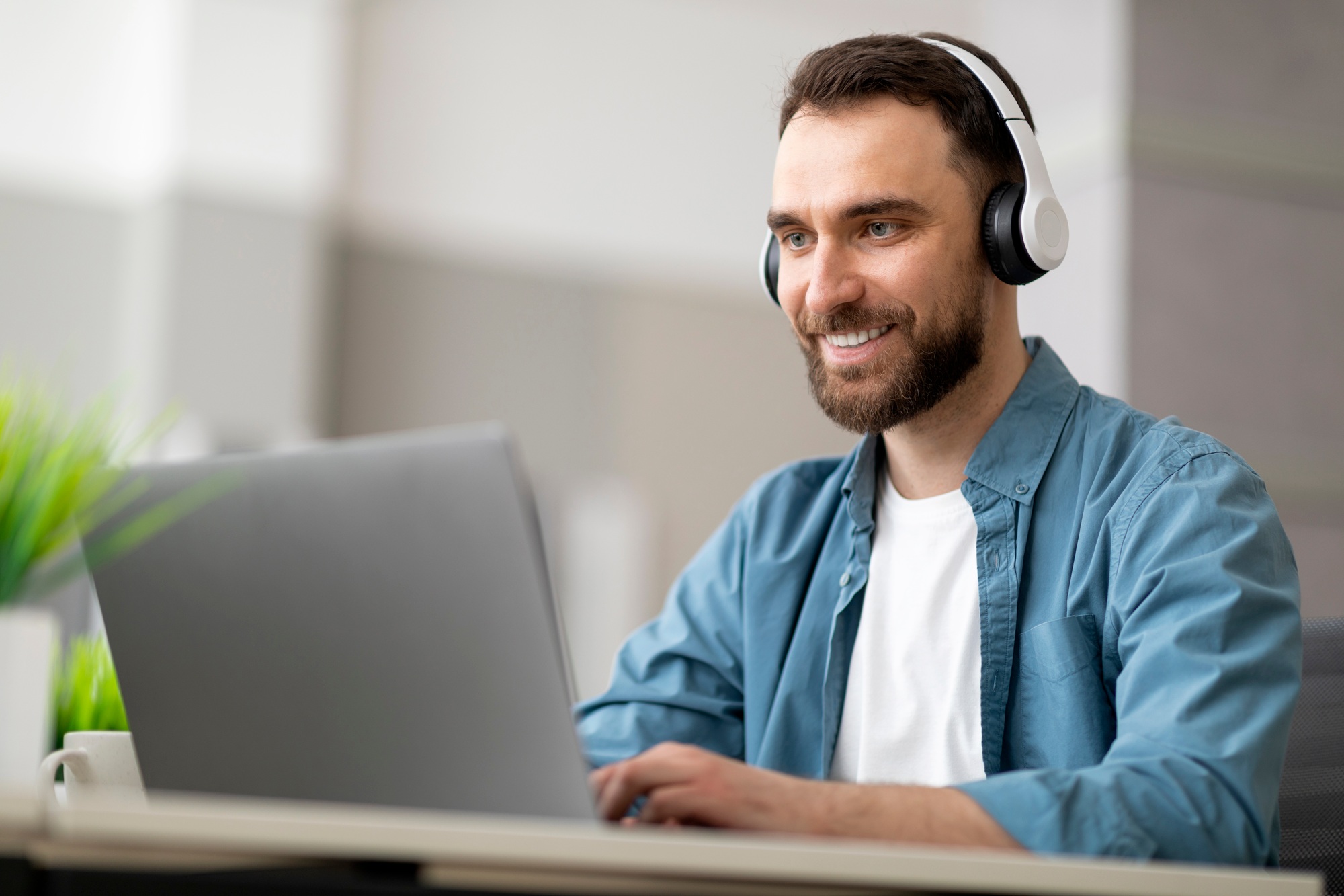 Smiling male employee wearing wireless headphones working on computer in modern office