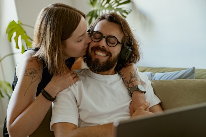 Smiling man working on laptop while wife is kissing him at home. Distance work concept