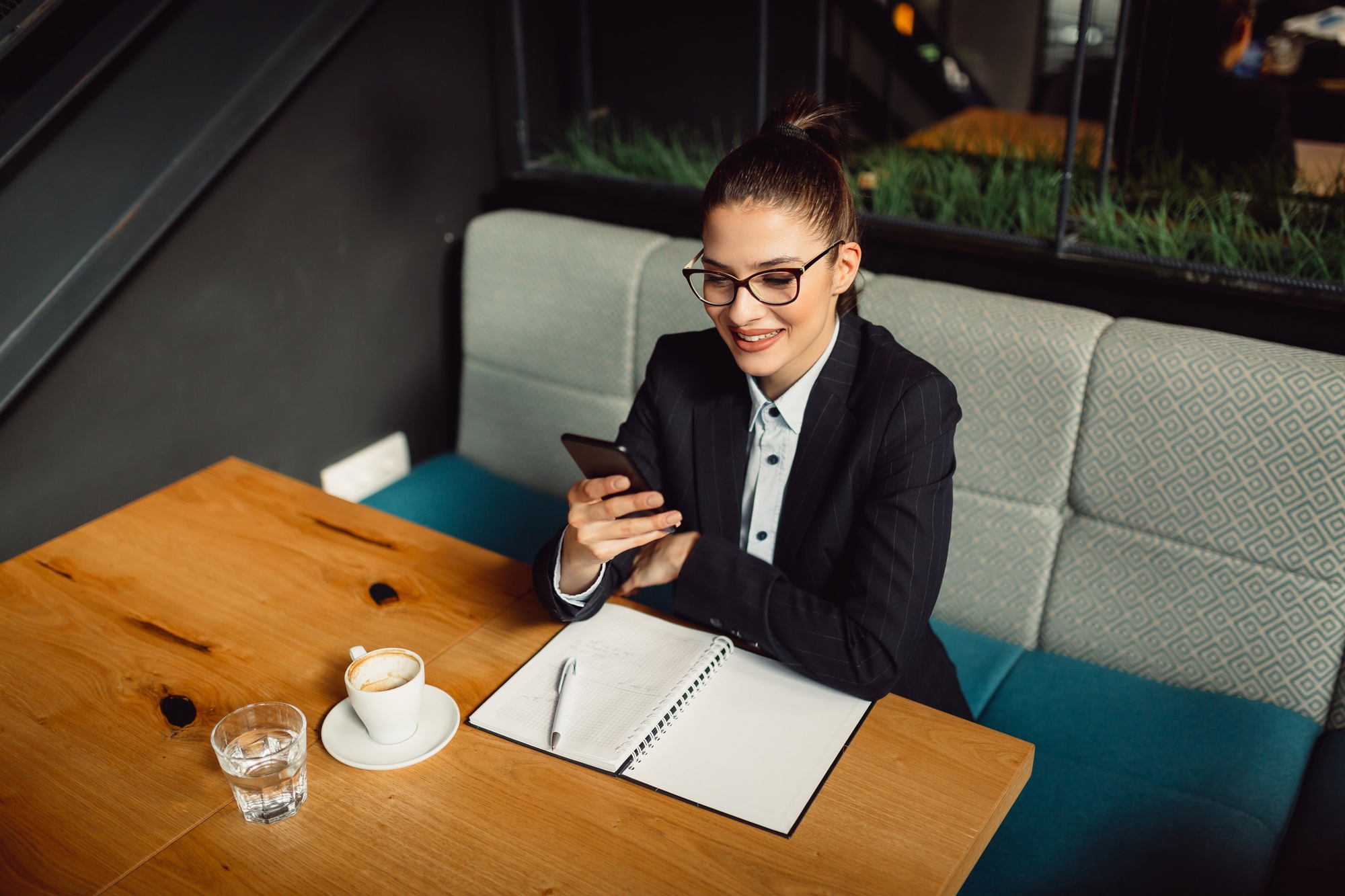 Thoughtful confident female in coffee shop wearing eyewear using mobile phone.