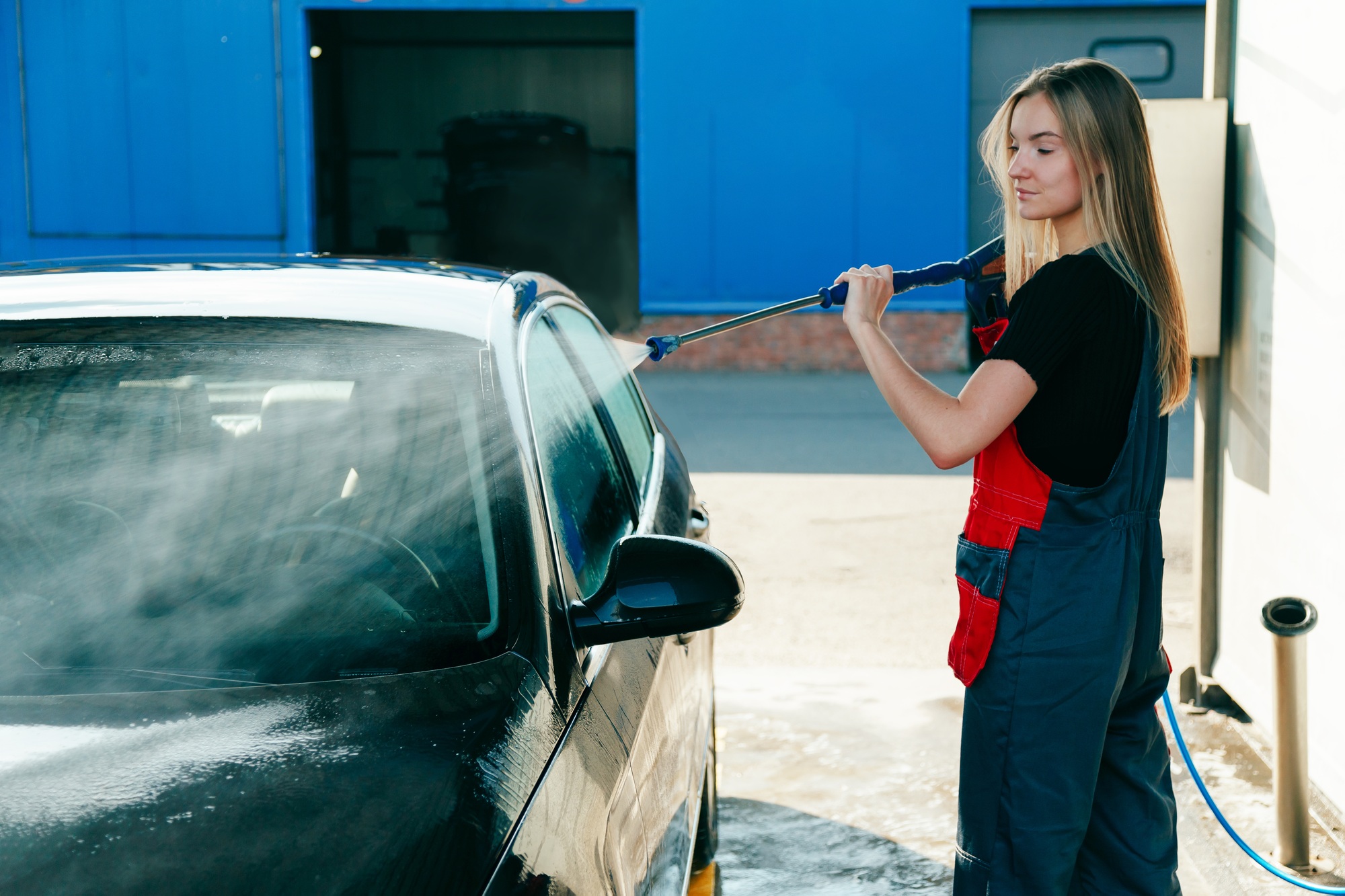 Woman Worker in Uniform Washing Car with High Pressure Gun with Hose at Car Wash Station