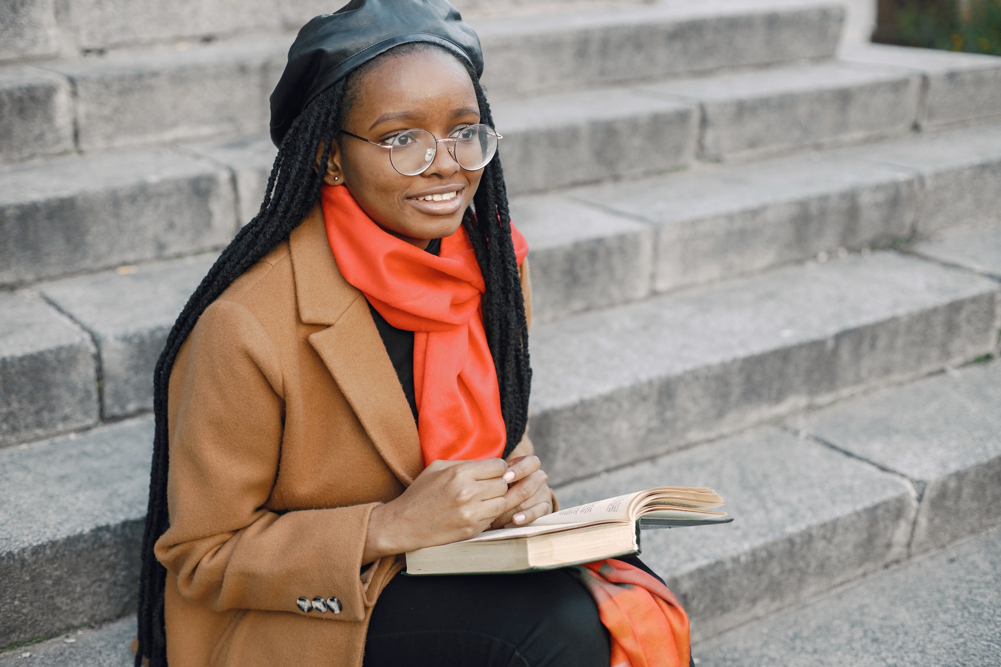 Young black woman in a coat sitting on a stairs and reading a book