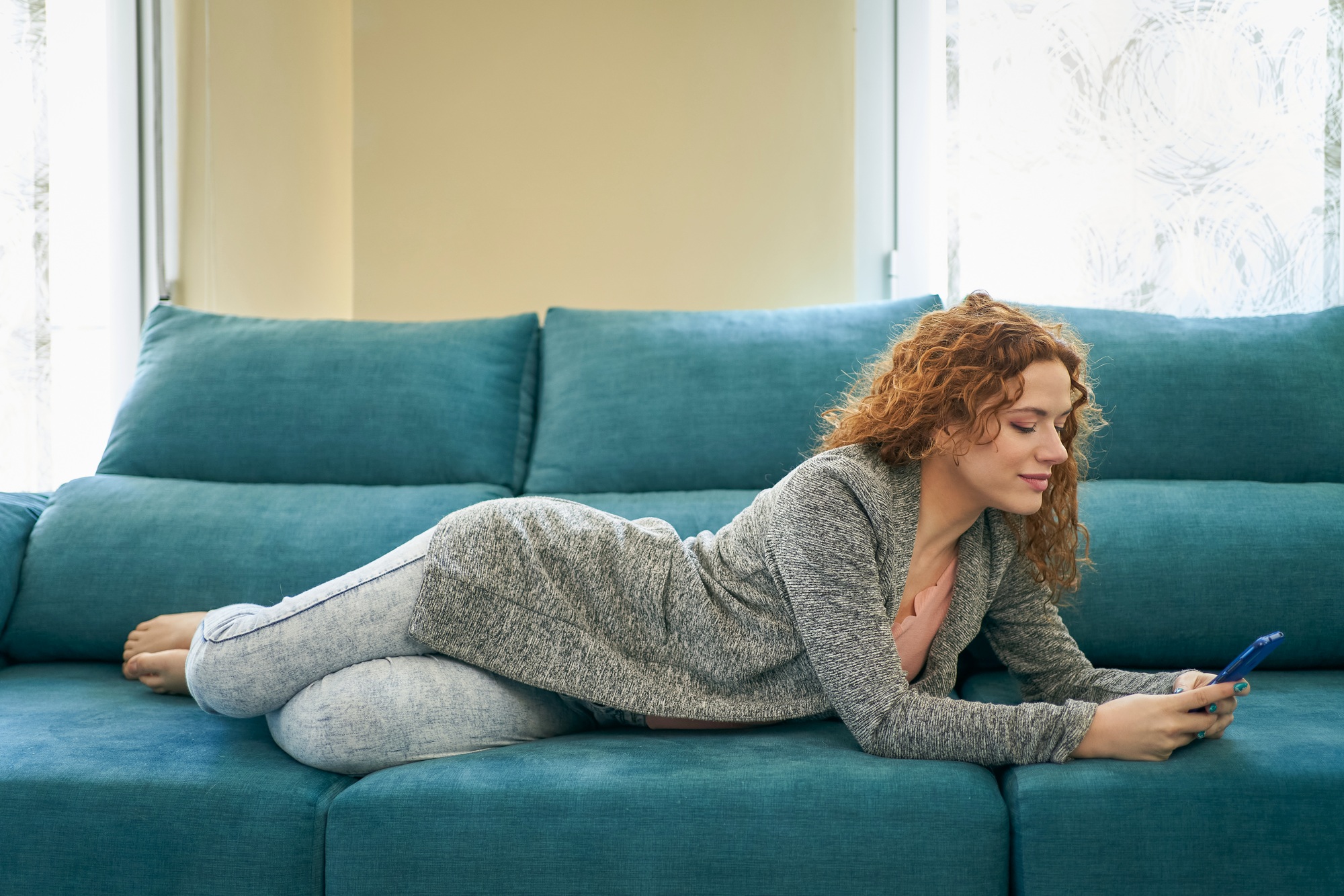 Young blogger publishing a new post on her website via her smartphone while lying on a sofa