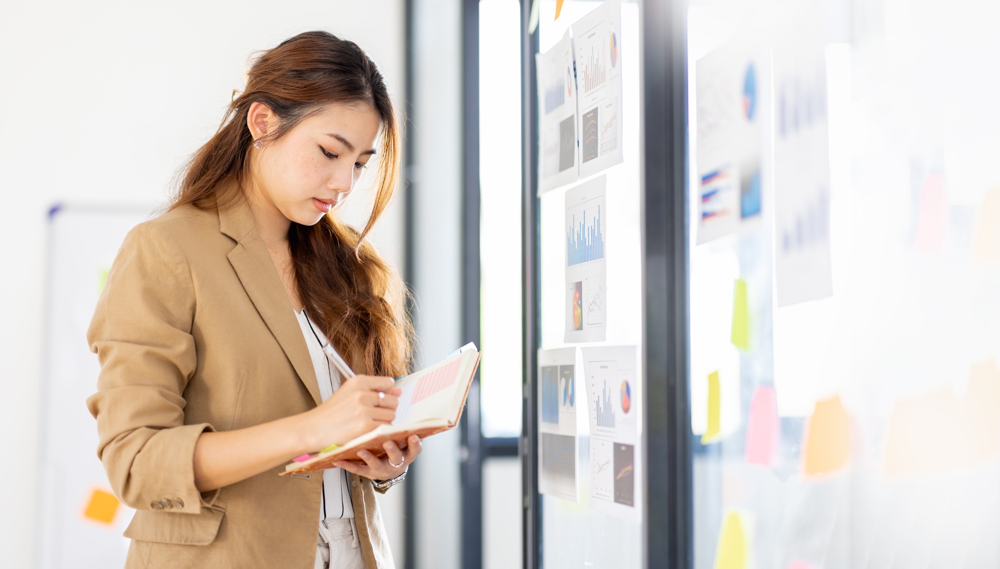 Young Business Asian woman using sticky notes and graph chart in glass wall analyzing strategy busin