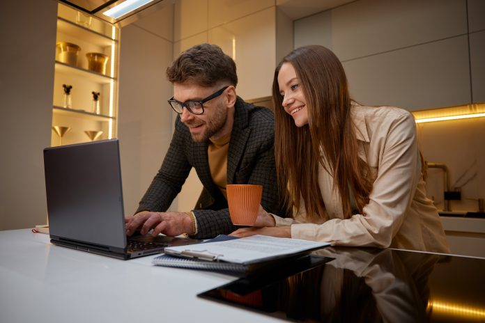 Young family using laptop on kitchen, husband helping loving wife with project