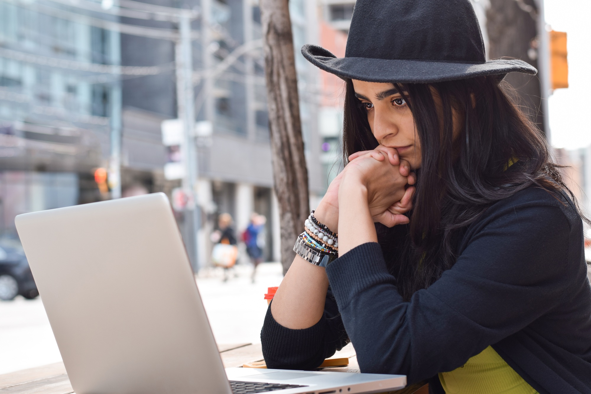 Young woman in fedora hat sitting at outdoor city street cafe using laptop computer