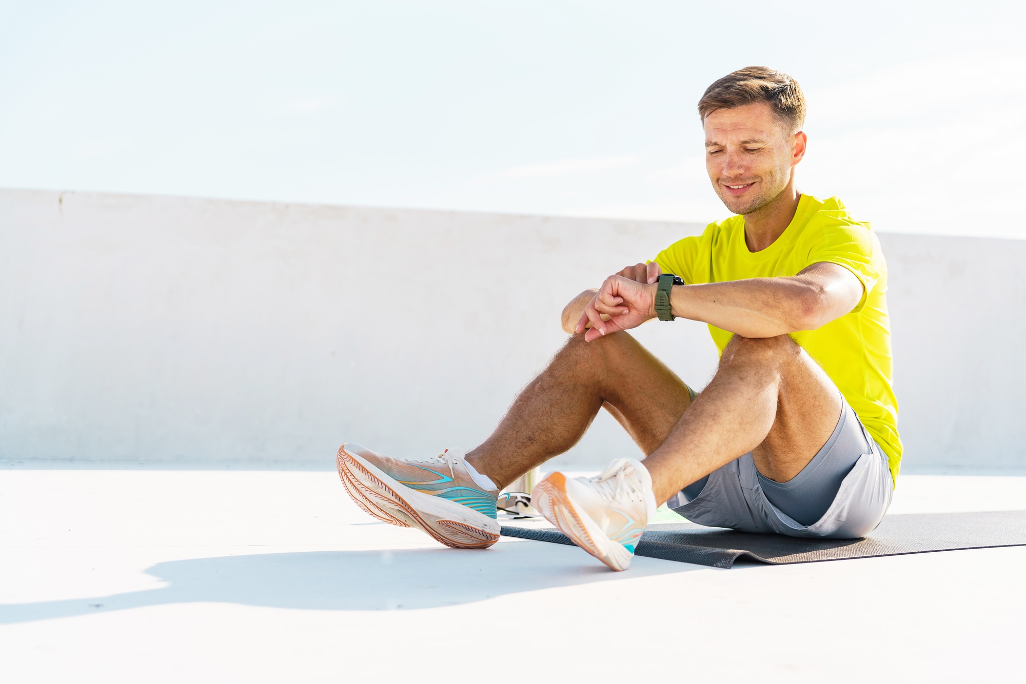 A man in a bright yellow shirt sits on the ground, checking his watch after a workout, relaxed