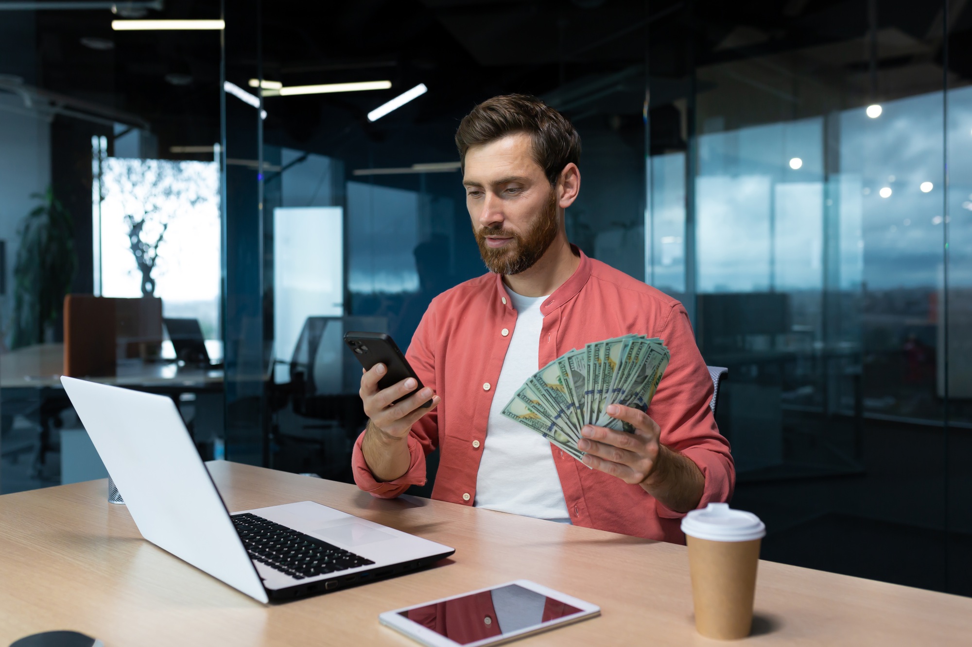 A young businessman, freelancer, banker sits in the office at a table with a laptop, holds cash