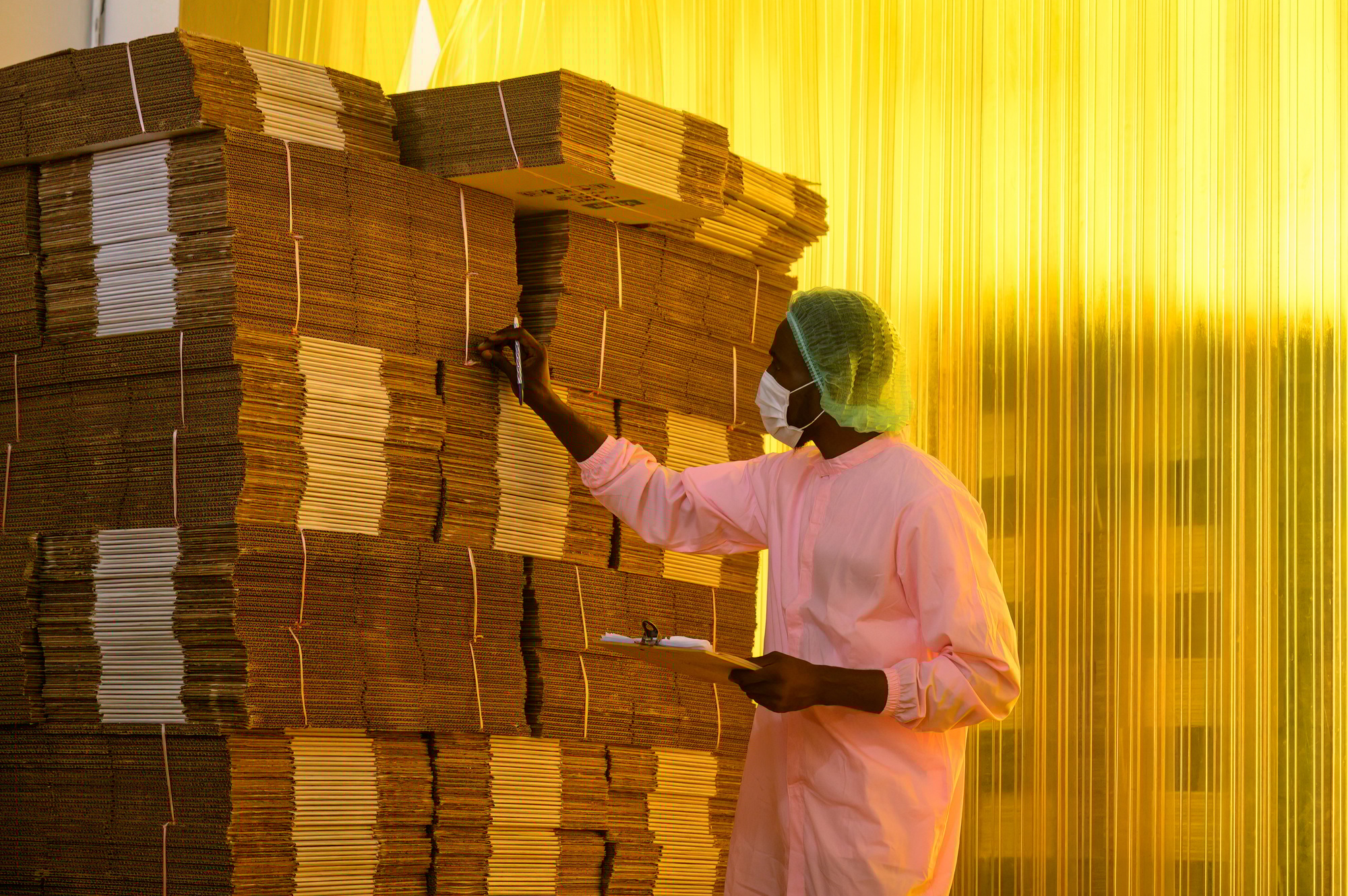 African male employee checking with clipboard at stacked cardboard package stuck in warehouse