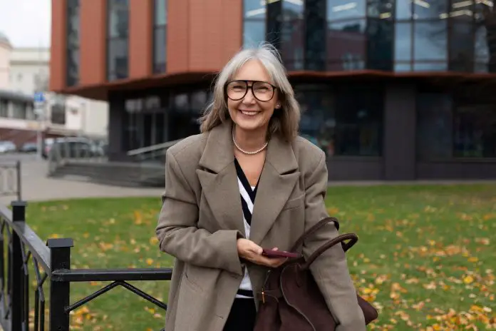 An older businesswoman smiles warmly while holding her phone and bag in front of a modern office