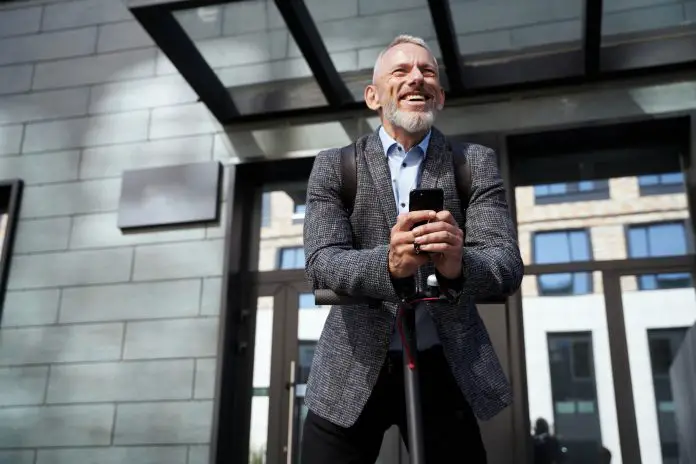 Cheerful middle aged businessman smiling, holding his mobile phone while standing with electric