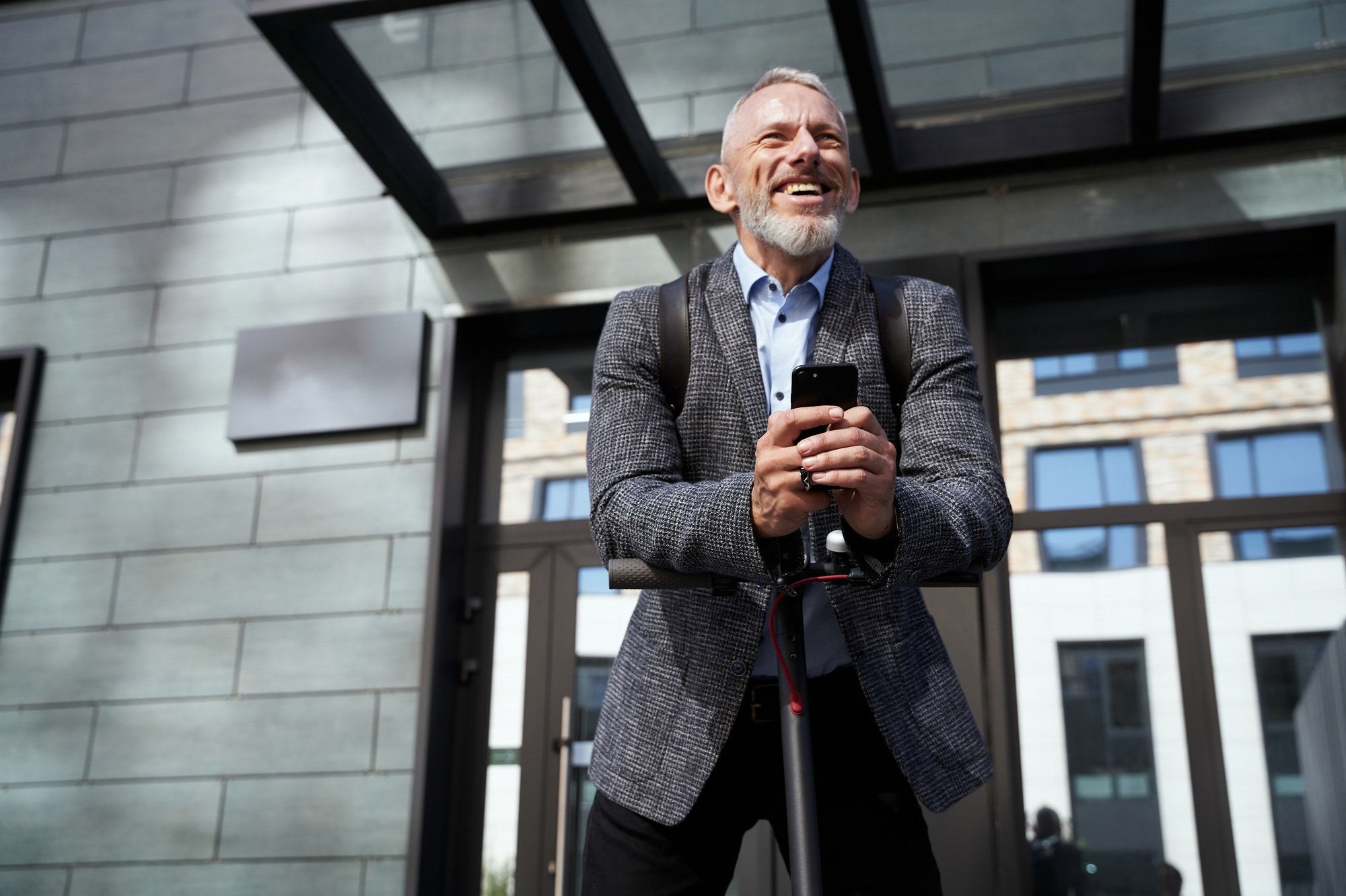 Cheerful middle aged businessman smiling, holding his mobile phone while standing with electric
