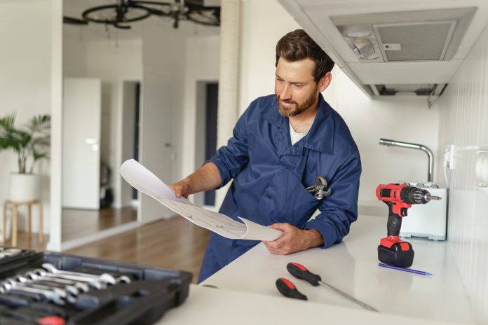 Handyman in uniform standing on kitchen with home plan and tool box before starting work