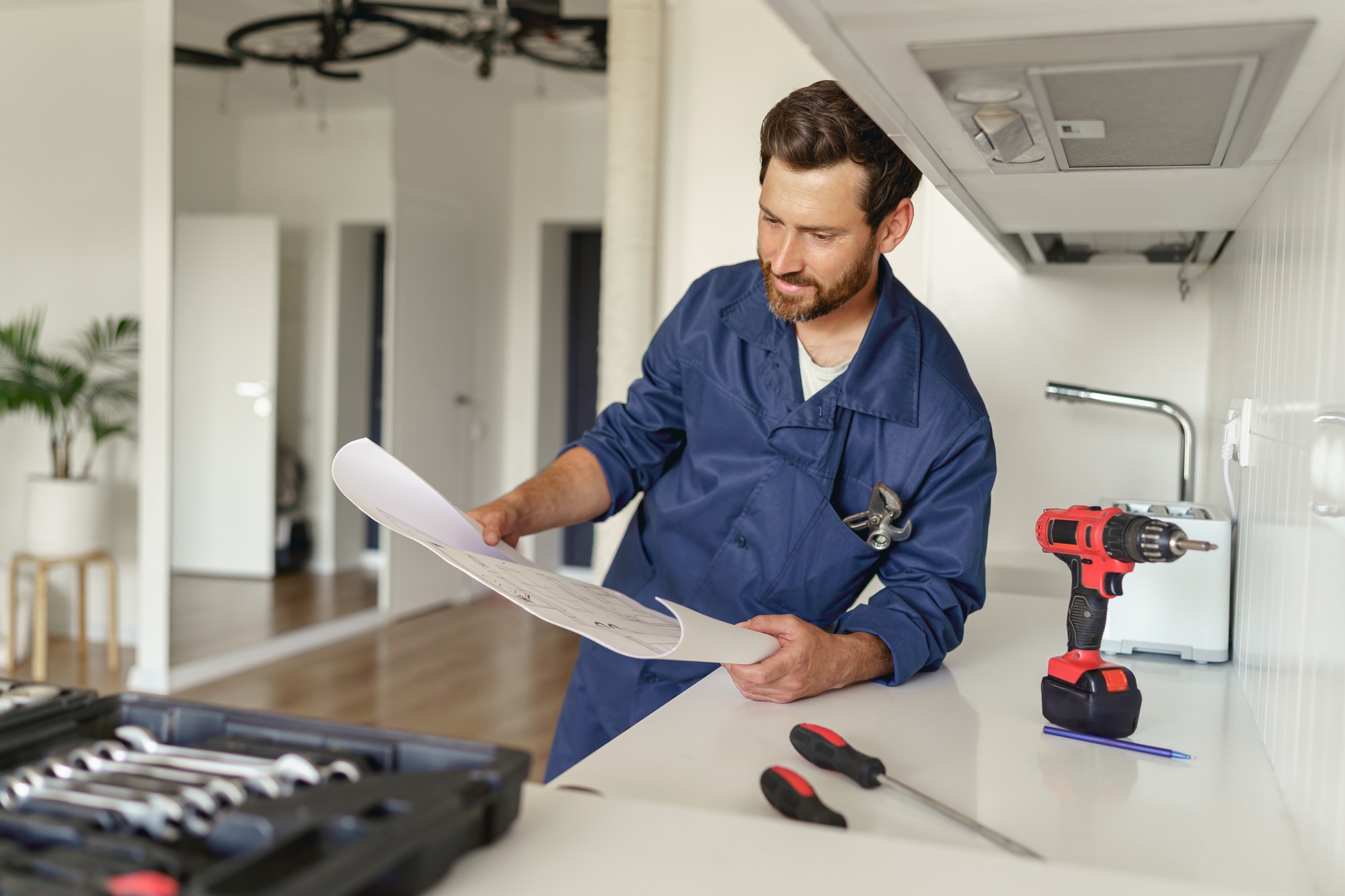 Handyman in uniform standing on kitchen with home plan and tool box before starting work