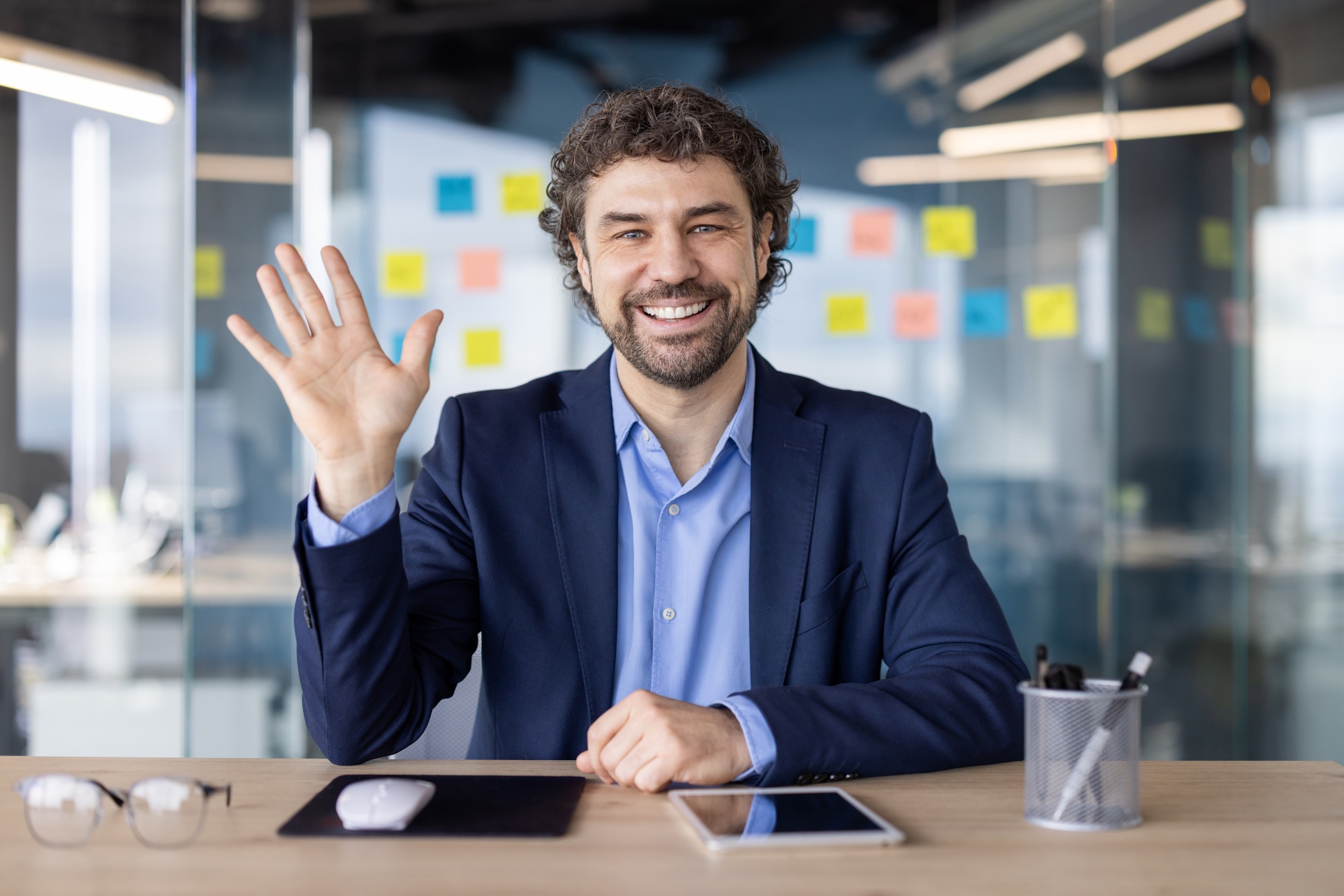 Man in business attire waves at the camera during a video call in a modern office setting, smiling