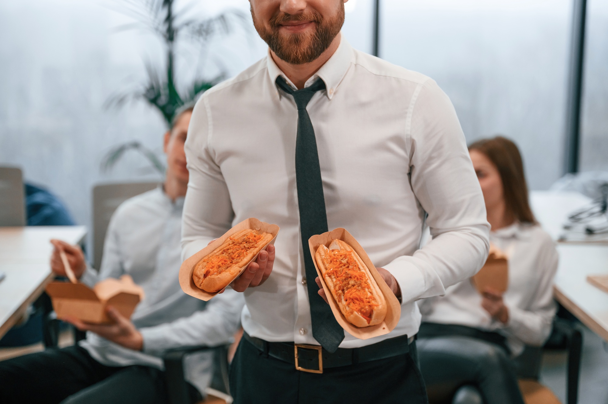 Man in formal wear is in the office, holding hot dogs in hands